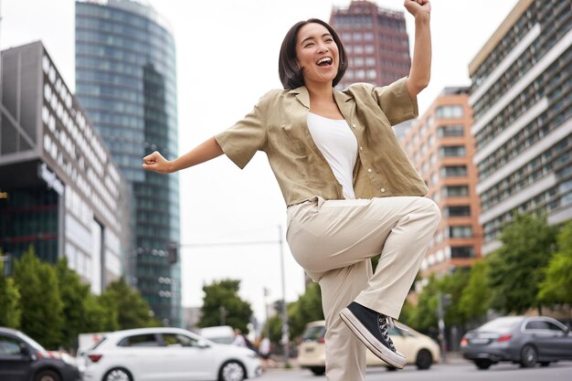 Portrait of dancing asian woman raising hands up and feeling happy posing in city triumphing feeling