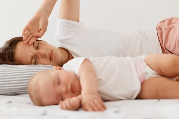 Portrait of dark haired female wearing white casual t shirt lying on bed with little baby daughter, posing indoor, woman looking at her infant girl with tired expression.