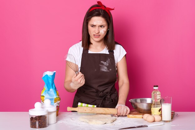 Portrait of dark haired girl in apron soiled with flour, t shirt and red hair band, stands with whisk in hands and feels disgusted from baking pies, wants to have rest. Baker makes delicious cookies.