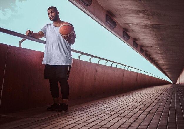 Portrait of a dark-skinned bearded guy dressed in a white shirt and sports shorts leaning on a guardrail while standing with basketball.