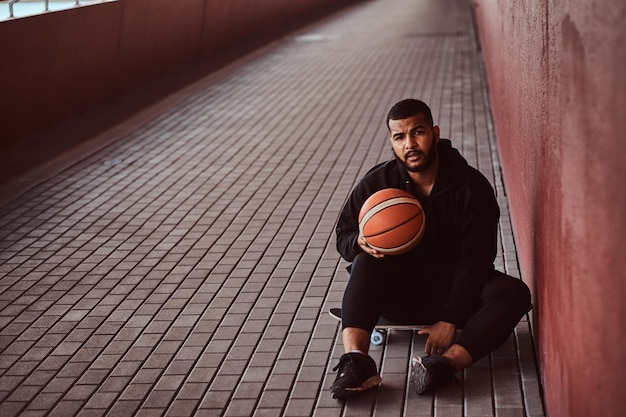 Il ritratto di un ragazzo dalla pelle scura vestito con una felpa con cappuccio nera e pantaloncini sportivi tiene un pallone da basket mentre è seduto su uno skateboard e si appoggia a un muro.