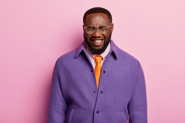 Portrait of dark skinned male employee has hysterical laugh, positive mood, smiles broadly, wears purple jacket, orange tie