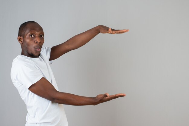 Portrait of dark skinned man in white t-shirt holding open space on gray wall