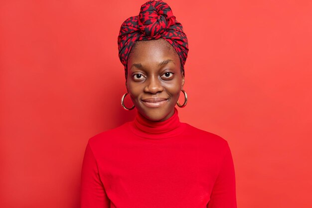 Portrait of dark skinned young lady smiles happily looks directly at camera wears kerchief on heaad poloneck isolated on bright vivid red.inheritancee expressions concept. Monochrome