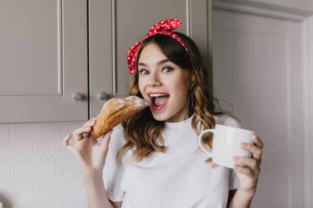 Портрет молодой женщины debonair есть круассан с удивленной улыбкой. Элегантная кудрявая девушка позирует на уютной кухне с чашкой чая.