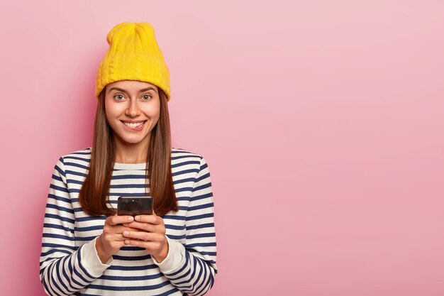 Portrait of delighted cheerful woman bites lips and looks happily at camera, uses modern cell phone, dressed in stylish clothes poses over rosy background