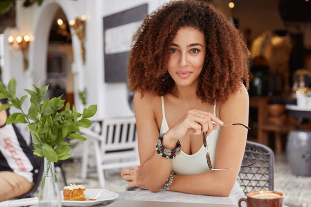 Portrait of delighted restful young female with bushy hairdo and dark skin, holds shades, enjoys tasty dessert in restaurant has informal meeting with partner during spare time.