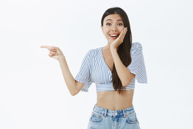 Portrait of delighted and surprised emotive touched girlfriend with cropped blouse touching cheek amazed and pleased pointing left