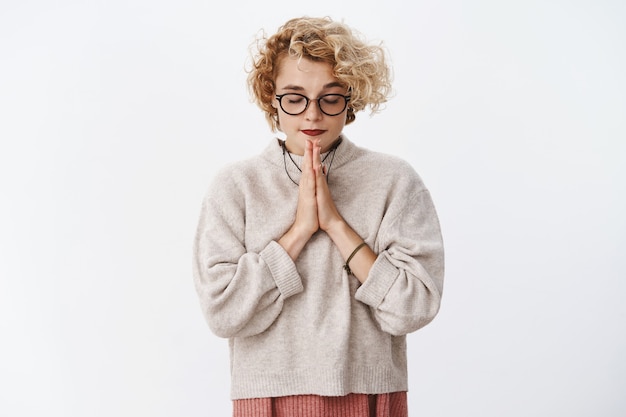Portrait of determined attractive stylish hipster woman with short curly haircut in glasses and sweater close eyes focus, holding hands in pray while making wish over white wall