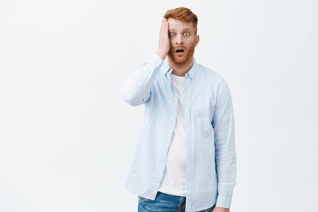 Portrait of devastated stunned mature redhead male in shirt, holding palm on face and dropping jaw from shook