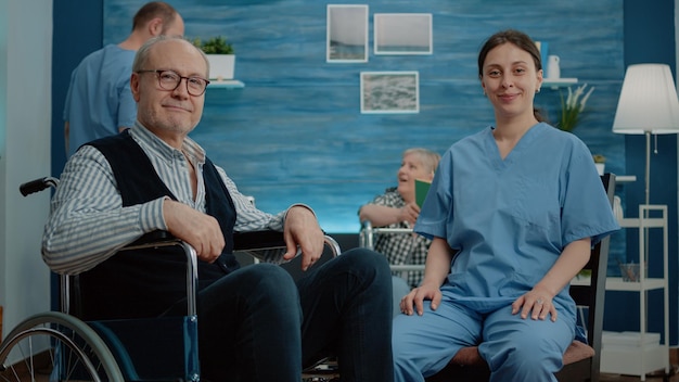 Portrait of disabled senior man sitting with woman nurse in nursing home, looking at camera. Retired adult in wheelchair getting help and assistance from medical assistant at facility