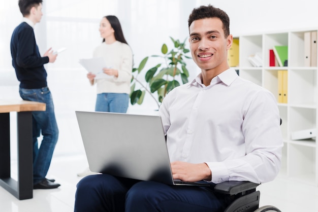 Portrait of a disabled smiling young businessman sitting on wheelchair with laptop and business colleague in the background