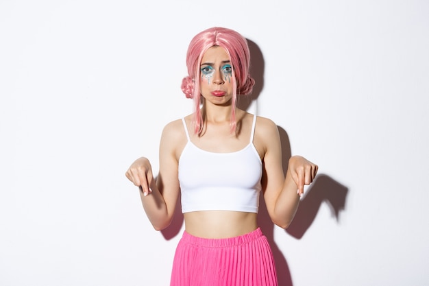 Portrait of disappointed cute sulking girl in pink wig and halloween outfit, looking upset while pointing fingers down, complaining, standing over white background