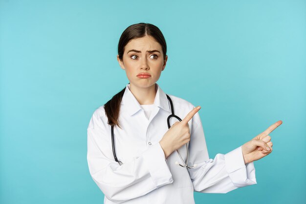 Portrait of disappointed doctor woman medical worker pointing fingers right and looking sad regret g...