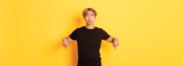 Портрет разочарованного мрачного азиатского парня, который расстроен и показывает пальцами вниз с сожалением и разочарованием