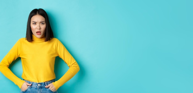 Free photo portrait of disappointed korean woman in yellow sweater frowning and looking upset standing over blu