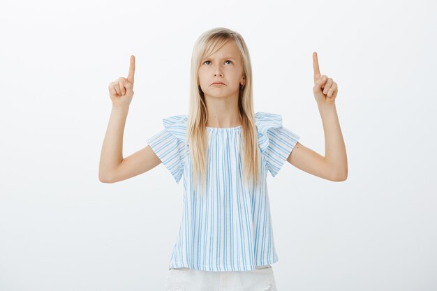 Portrait of disappointed offended cute blond girl in blue blouse, looking and pointing up with index fingers, frowning from displeased emotions, seeing person she dislikes over gray wall
