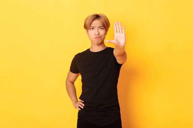 Portrait of disappointed serious asian man, smirking displeased and extend hand, showing stop gesture, yellow wall