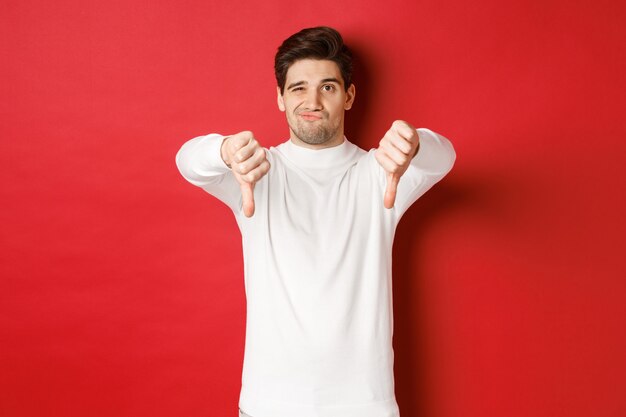 Portrait of disappointed skeptical guy wearing white sweater showing thumbsdown to disagree express ...