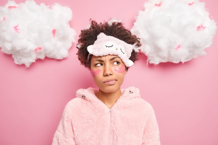 Free Photo portrait of discontent thoughtful curly woman looks pensively aside smirks face applies patches under eyes wears sleepmask nightwear isolated over pink wall