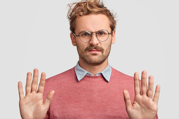 Free photo portrait of discontent young man with grumpy expression, makes stop symbol, keeps palms in front, has negative facial expression