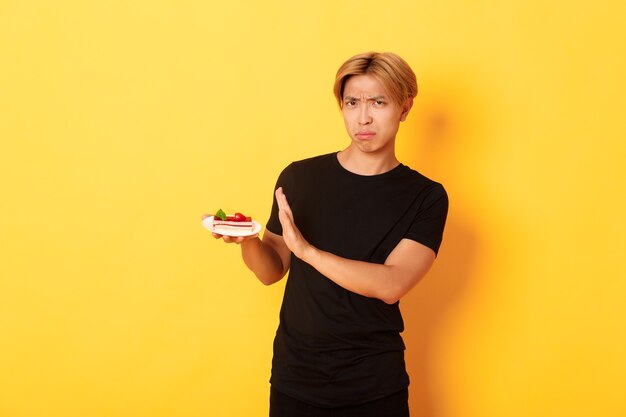 Портрет возмущенного и расстроенного молодого азиатского парня отказывается есть торт, выглядит недовольным и демонстрирует жест отказа, желтая стена.