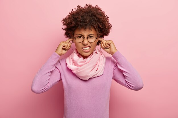 Portrait of displeased African American woman plugs ears and smirks face, avoids loud noise, wears transparent glasses, casual purple wear