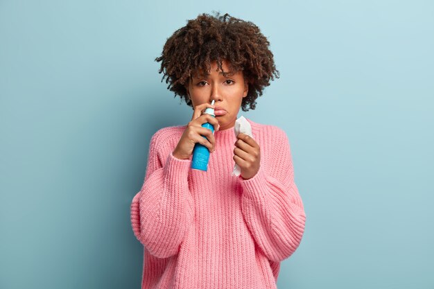 Portrait of displeased Afro American woman sniffs nasal aerosol, feels sick, has running nose, uses medication for blocked nose, holds tissue, has sad facial expression, wears pink jumper.