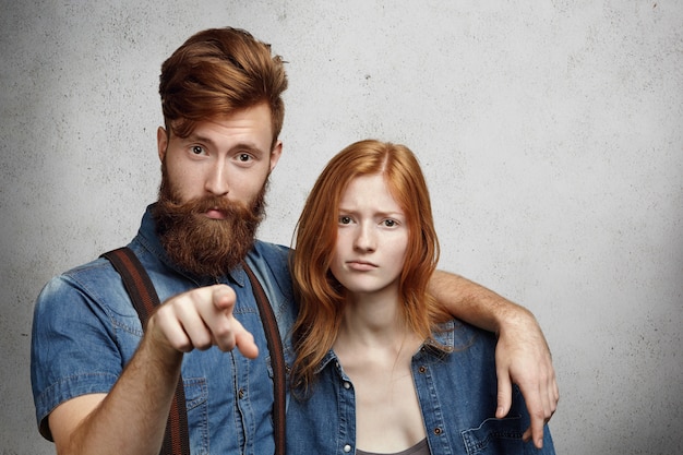 Portrait of displeased or angry man with stylish beard pointing  and hugging protectively beautiful redhead woman with hurt look, defending his girlfriend and warning you not to touch her