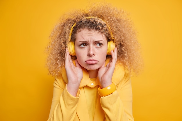 Free photo portrait of displeased curly haired young european woman keeps hands on headphones has frustrated face expression purses lips