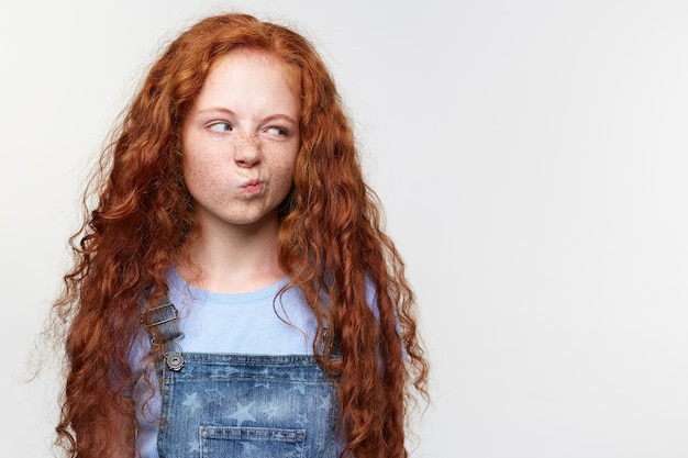 Free photo portrait of displeased cute freckles little girl with ginger hair, looks with disgust at the copy space on the left side, tands over white background.