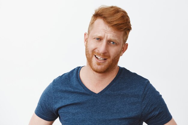 Portrait of displeased doubtful and disappointed handsome mature male with red hair, frowning and staring, expressing dislike or hesitation over grey wall