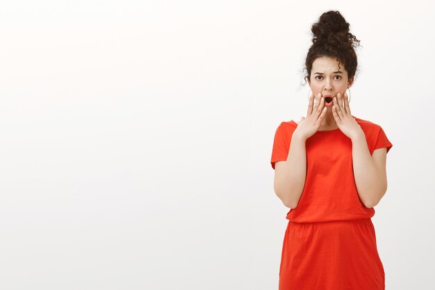 Portrait of displeased shocked feminine woman with curly hair in casual stylish dress, holding hand near opened mouth