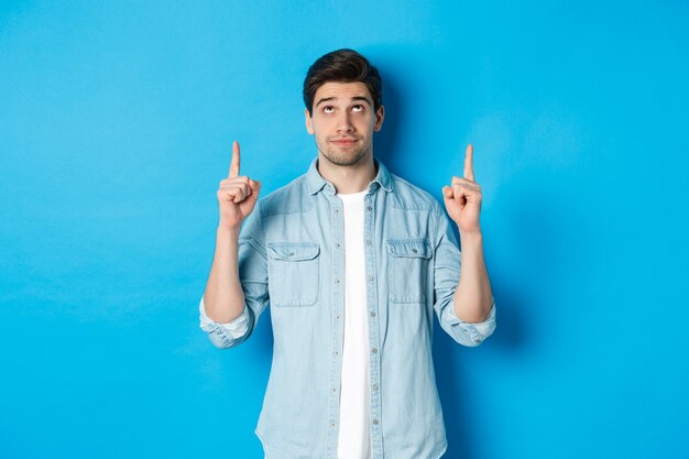 Portrait of displeased and skeptical male model pointing fingers up, looking at something unpleasant, standing against blue background.