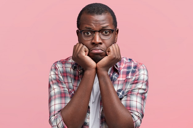 Portrait of displeased sorrorful dark skinned male curves lower lip and keeps hands under chin, dressed in casual shirt and eyewear