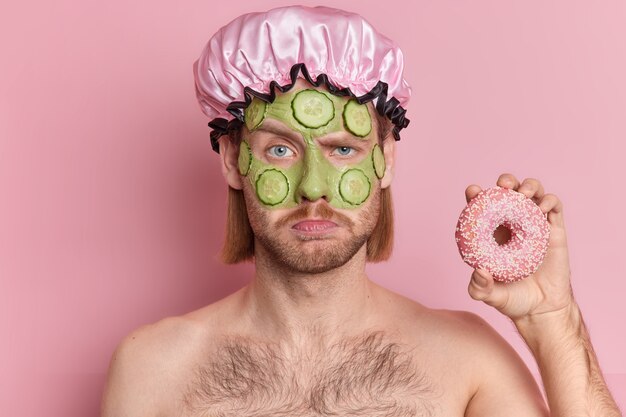 Portrait of displeased sullen man with mustache frowns face holds delicious donut stands topless indoor applies facial green mask cucumber slices for skin rejuvenation.