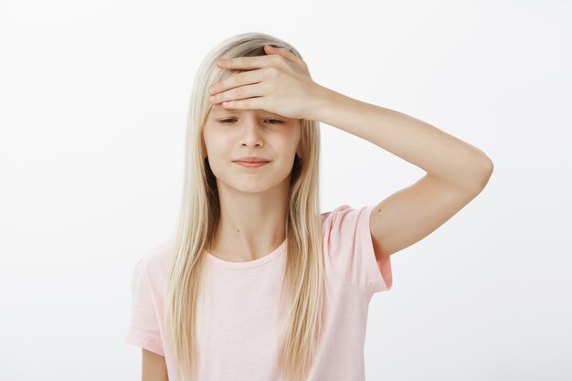 Portrait of displeased upset little girl with blond hair in pink t-shirt, holding palm on forehead and looking down with sad expression, feeling unwell and headache, standing with temperature