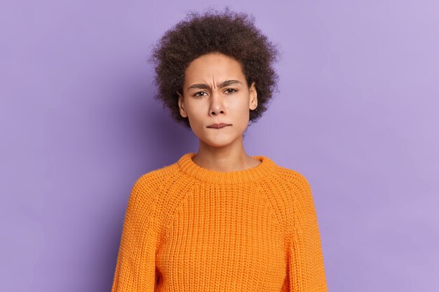Portrait of dissatisfied dark skinned girl with curly hair bites lips frowns face looks displeased dressed in knitted orange sweater.