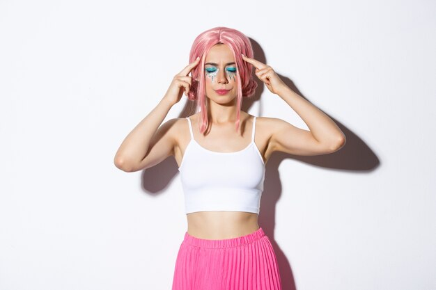 Portrait of distressed caucasian girl with pink wig and party makeup, standing with closed eyes and touching head