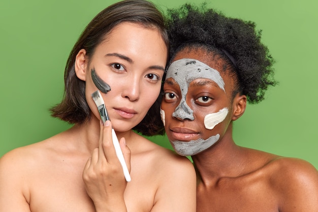 Portrait of diverse young women look seriously at camera apply clay mask use cosmetic brush stand topless