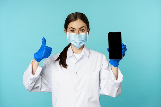 Portrait of doctor in medical face mask and gloves showing mobile phone app smartphone screen and th...