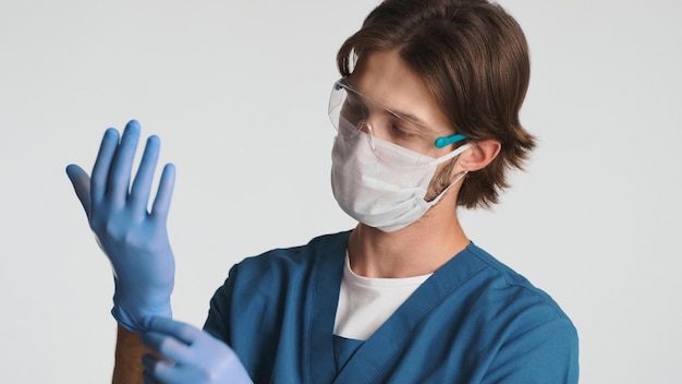 病院での就業日の準備をしている医療マスクと手袋を着用した医師の肖像画白い背景の上に立っている制服と保護された眼鏡に身を包んだ若いインターン