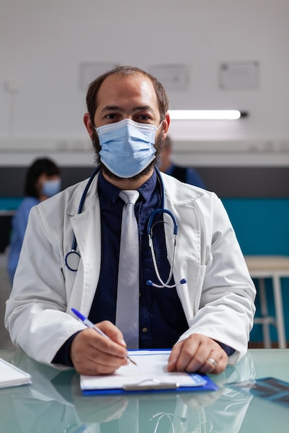 Portrait of doctor wearing white coat and face mask in cabinet, using clipboard paper and pen to take notes at checkup visit. Confident medic sitting at office desk during covid 19 pandemic.