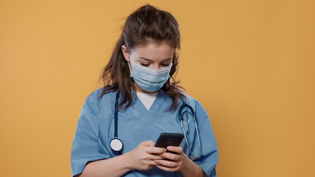 Portrait of doctor with stethoscope holding smartphone and texting showing green screen mockup at camera. Medic in hospital uniform wearing surgical mask using touchscreen device presenting copy space