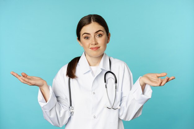 의사의 초상화, 어깨를 으쓱하는 여성 인턴, 웃으면서 미안하다고 말하고, 몰라요, 무지하고, 파란색 배경 위에 서 있습니다.