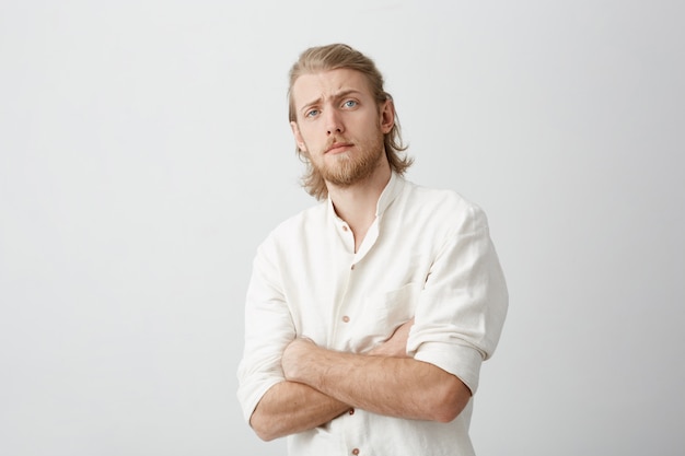 Portrait of doubtful attractive blond bearded man bending backwards, standing with crossed hands, lifting eyebrows and looking with disbelief