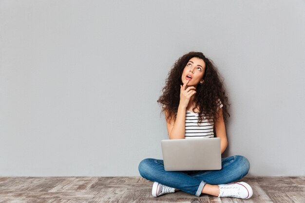 Portrait of dreaming female in casual clothes sitting with legs crossed on the floor with face upward working in silver computer over grey wall