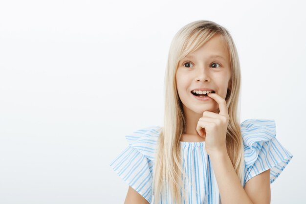 Portrait of dreamy adorable child with blond hair, looking aside and biting finger with broad smile, imaging