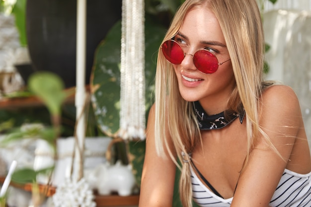 Free photo portrait of dreamy adorable woman with blonde hair slightly tied, wears red sunglasses