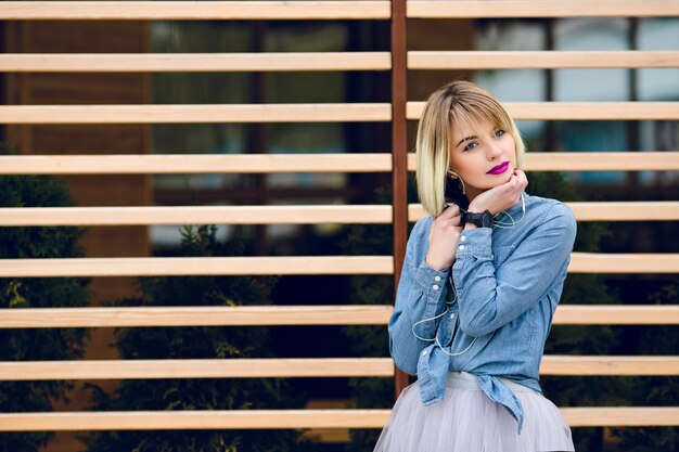 A portrait of a dreamy blonde girl with bright pink lips and nude makeup listening to music on a smartphone with striped wooden balks behind  